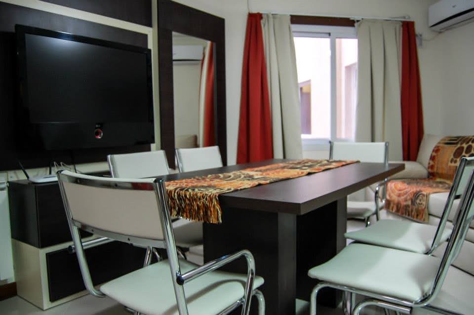 Comedor luminoso con mesa para seis, televisor grande y ventana, en un departamento de alquiler temporario en Villa Carlos Paz